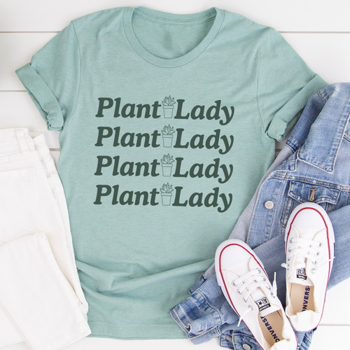 Plant Lady Tee shown in teal green with green words and a pair of jeans and tennis shoes sitting next to it on a white deck