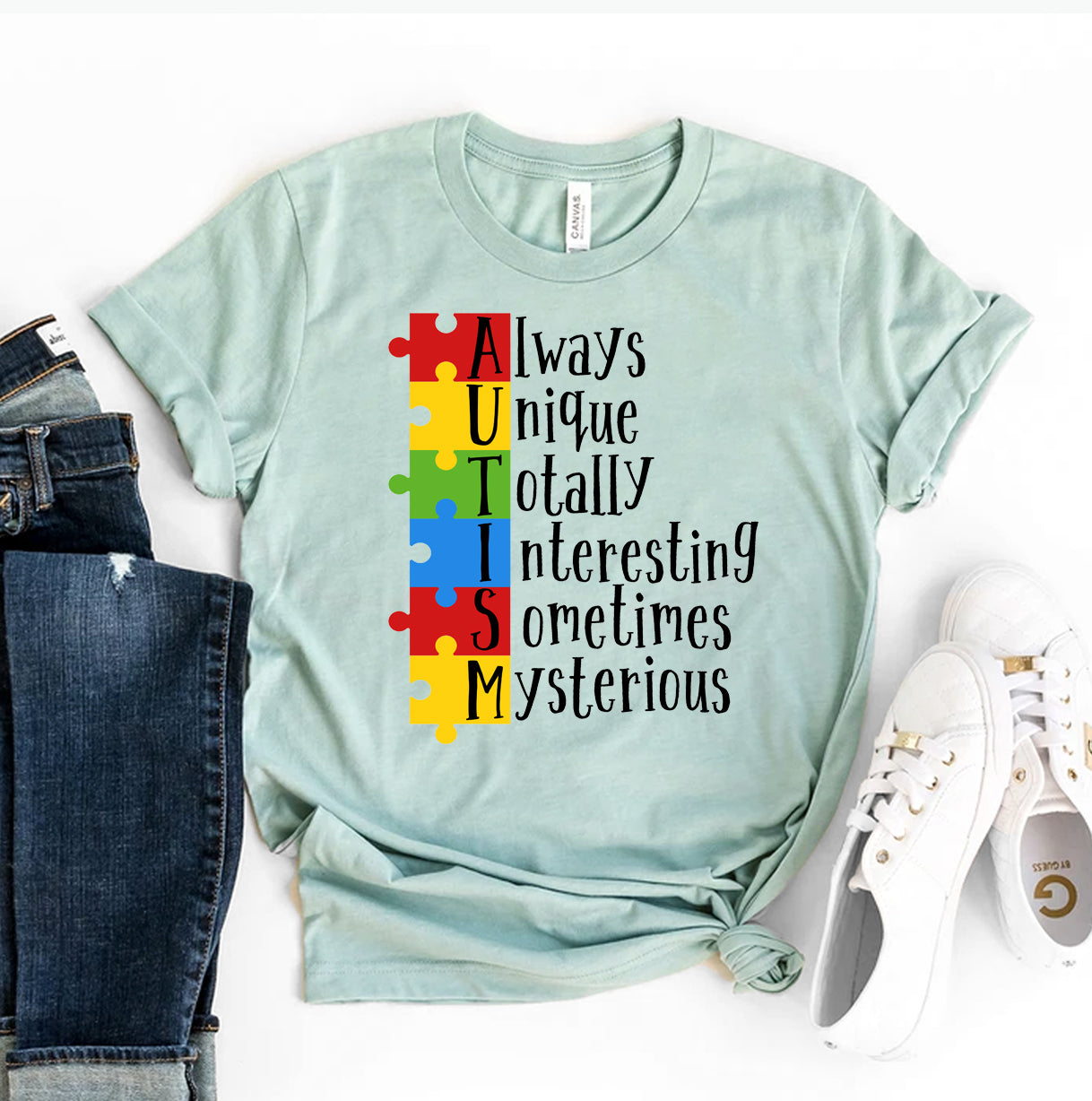 An Autism T-shirt shown with puzzle pieces arranged vertically with words next to them reading Always Unique Totally Interesting Sometimes Mysterious in teal green with a pair of jeans sitting next to it and a pair of white shoes