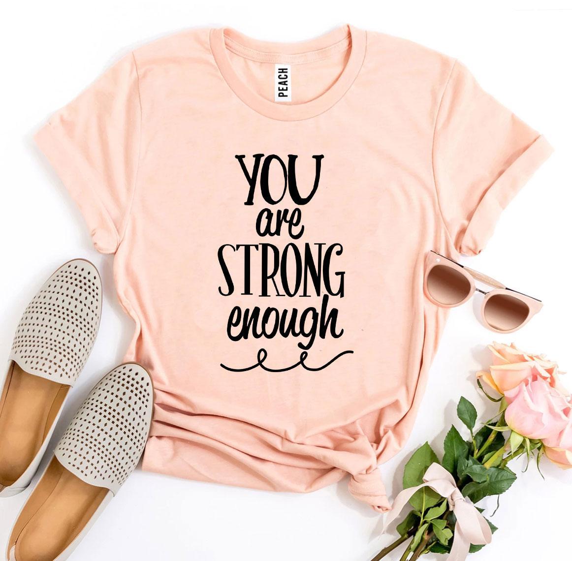 A peach You Are Strong Enough T-shirt shown with a pair of peach shades and tan slippers sitting next to it