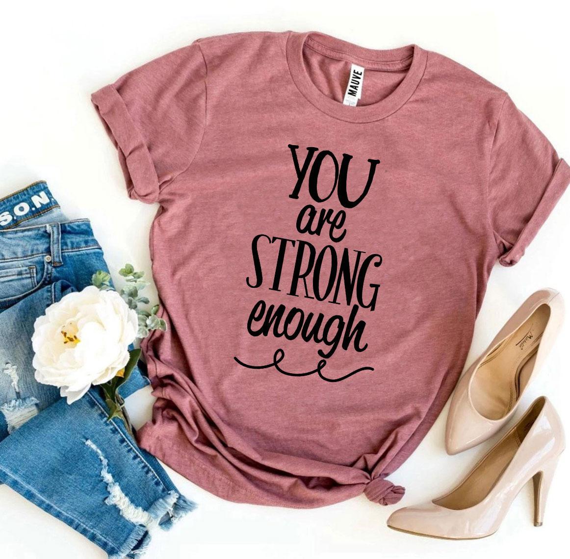 A dusty rose pink You Are Strong Enough T-shirt shown with a pair of tan heels and jeans sitting next to it