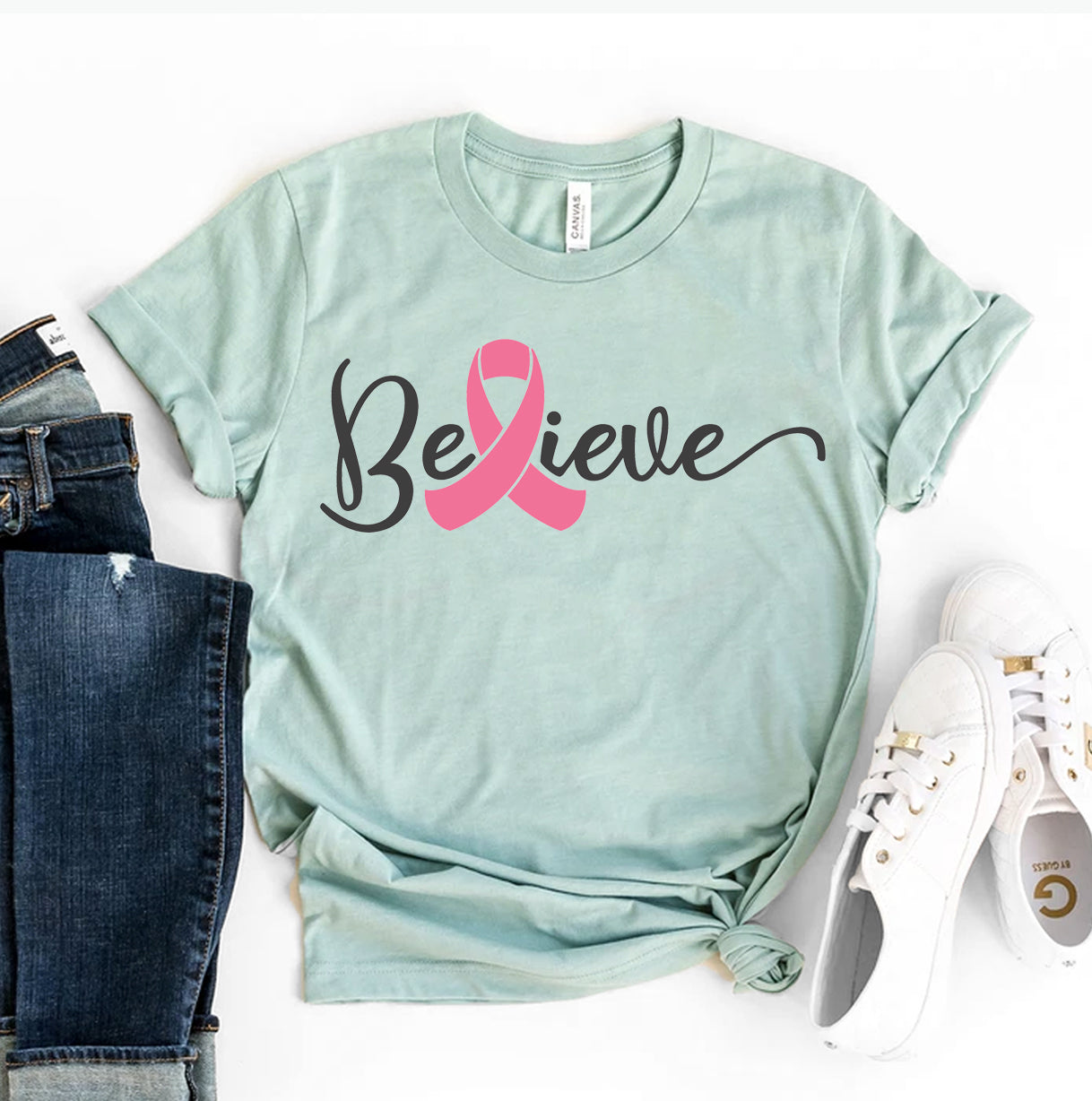 A Believe T-shirt shown in teal green and pink with a pair of jeans and white shoes sitting next to it