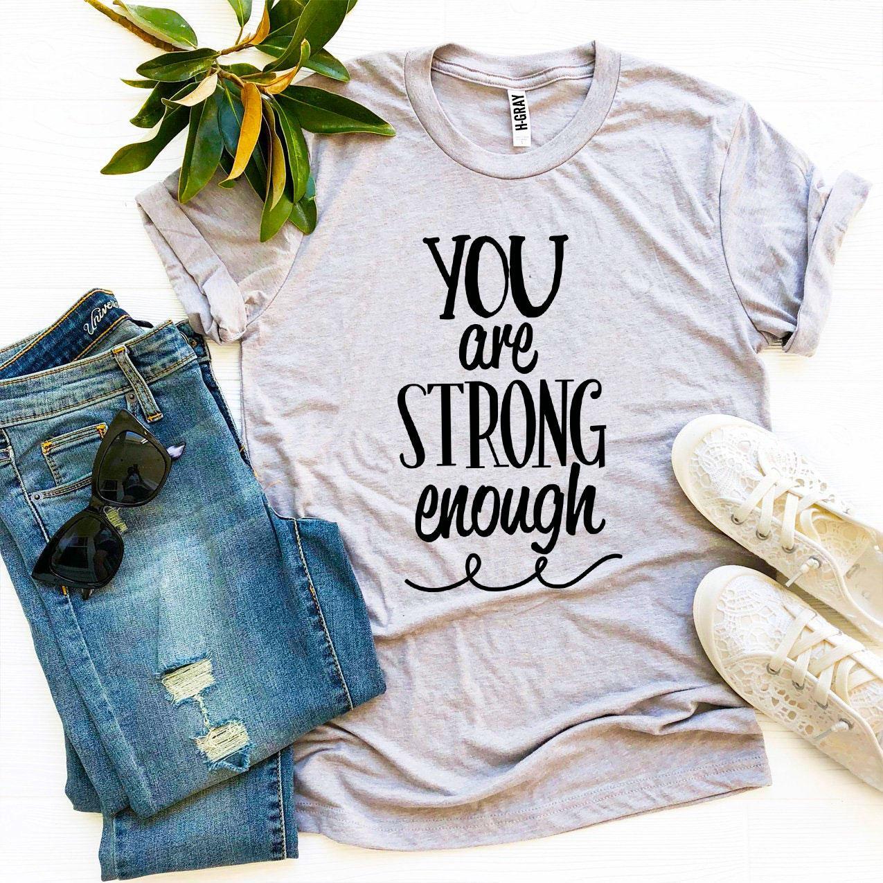 A grey You Are Strong Enough T-shirt with a pair of blue jeans sitting next to it and white tennis shoes