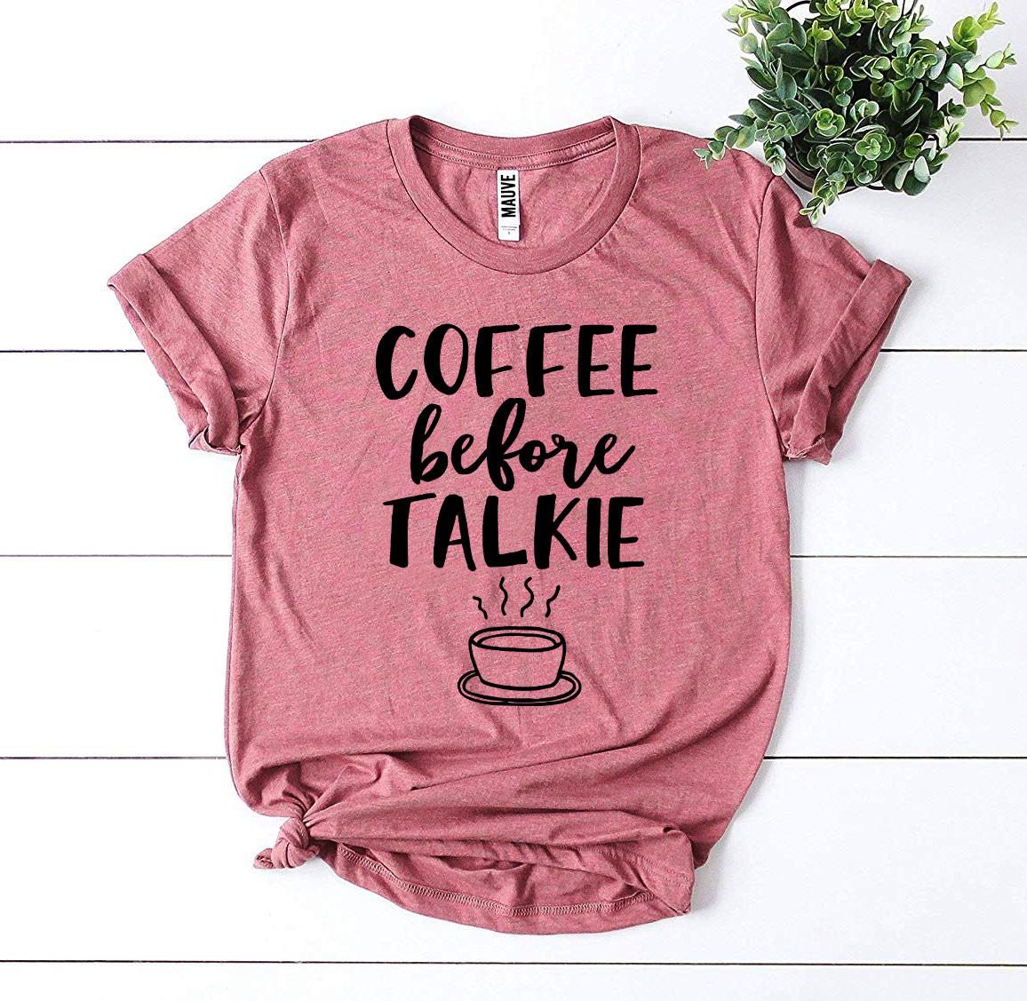 Pink Coffee Before Talkie T-shirt shown sitting on ship lap board with a green plant next to it