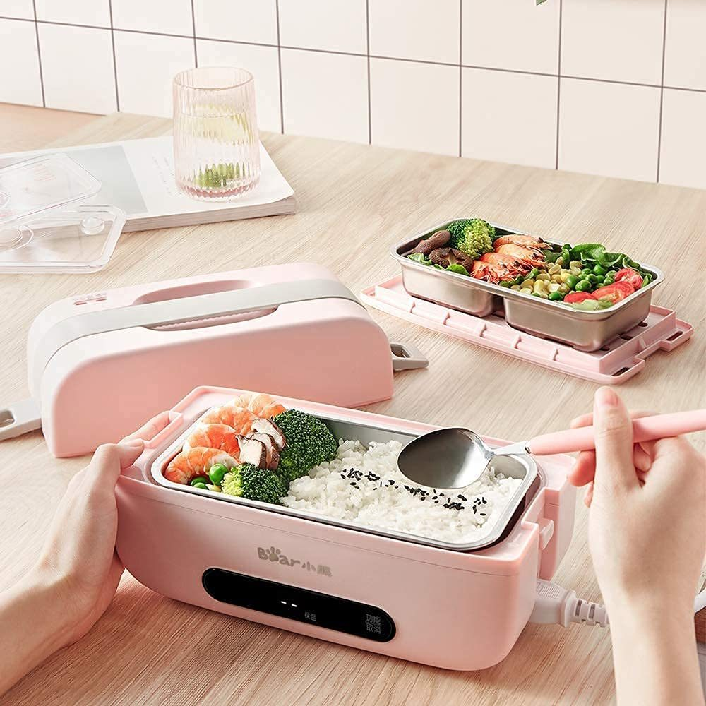 Pink self heating lunch box  sitting on a wooden table with a person eating the rice and veggies within