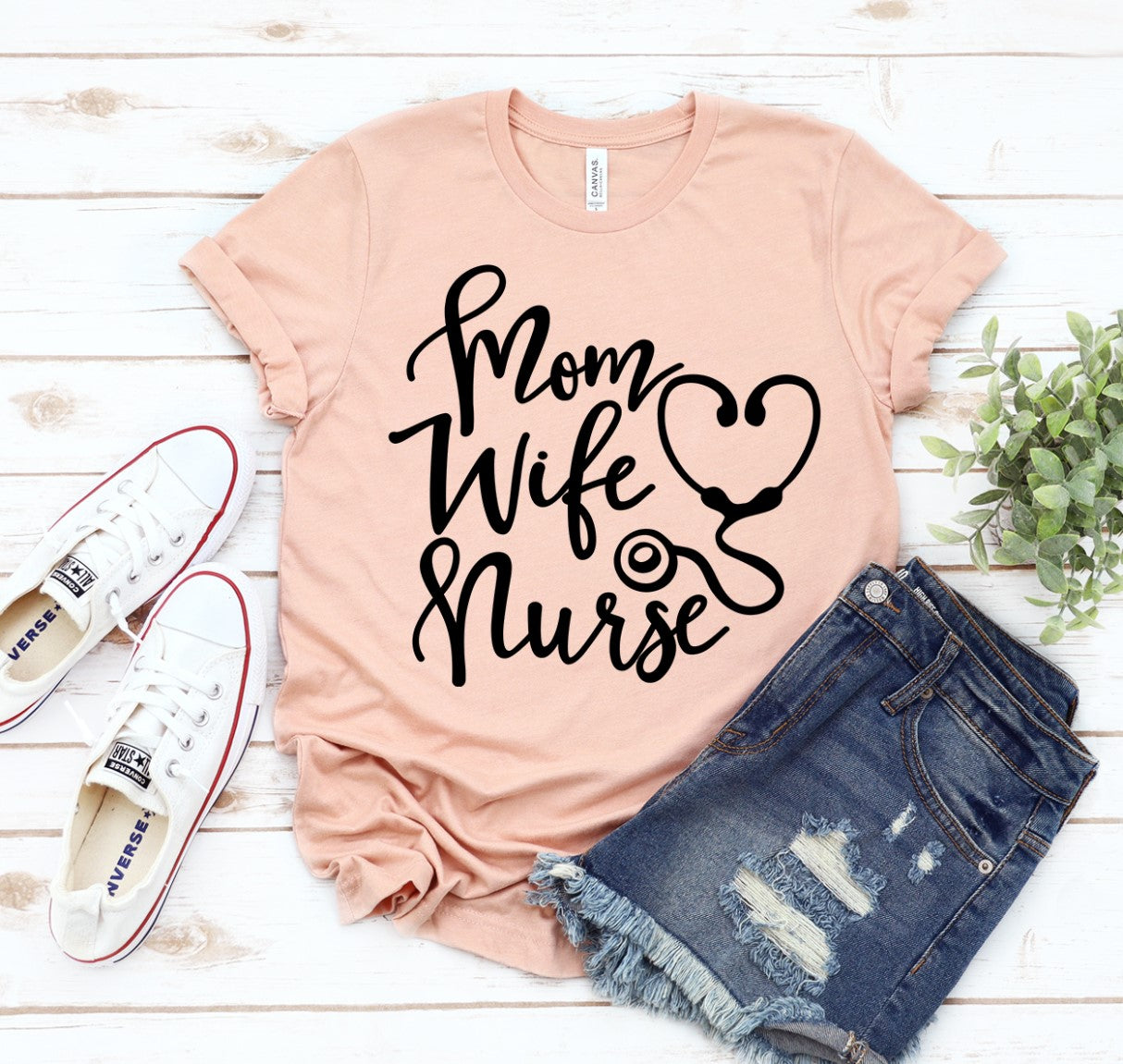 A Mom Wife Nurse T-shirt shown in peach sitting on white wooden deck with jeans and a pair of white tennis shoes sitting beside it